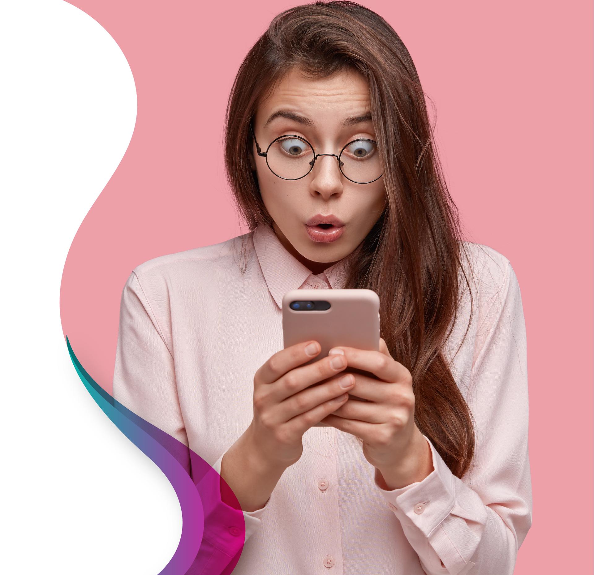 A young woman with long brown hair and round glasses looking at her pink smartphone with a shocked, wide-eyed expression against a pink background.